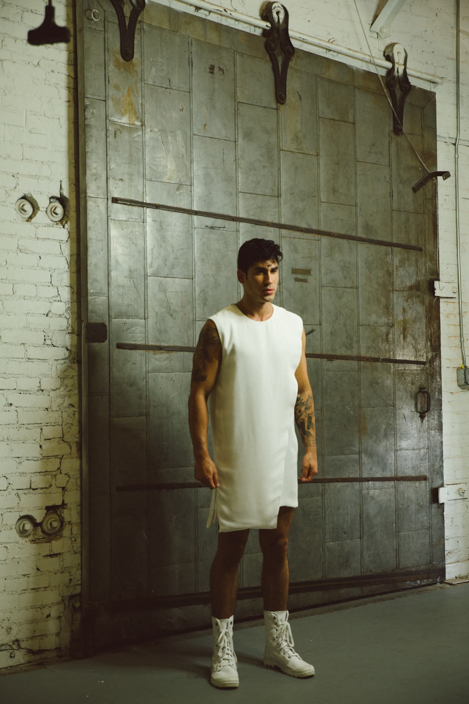 Male model in a sleeveless white extended tunic and white high-tops, industrial metal door behind him.